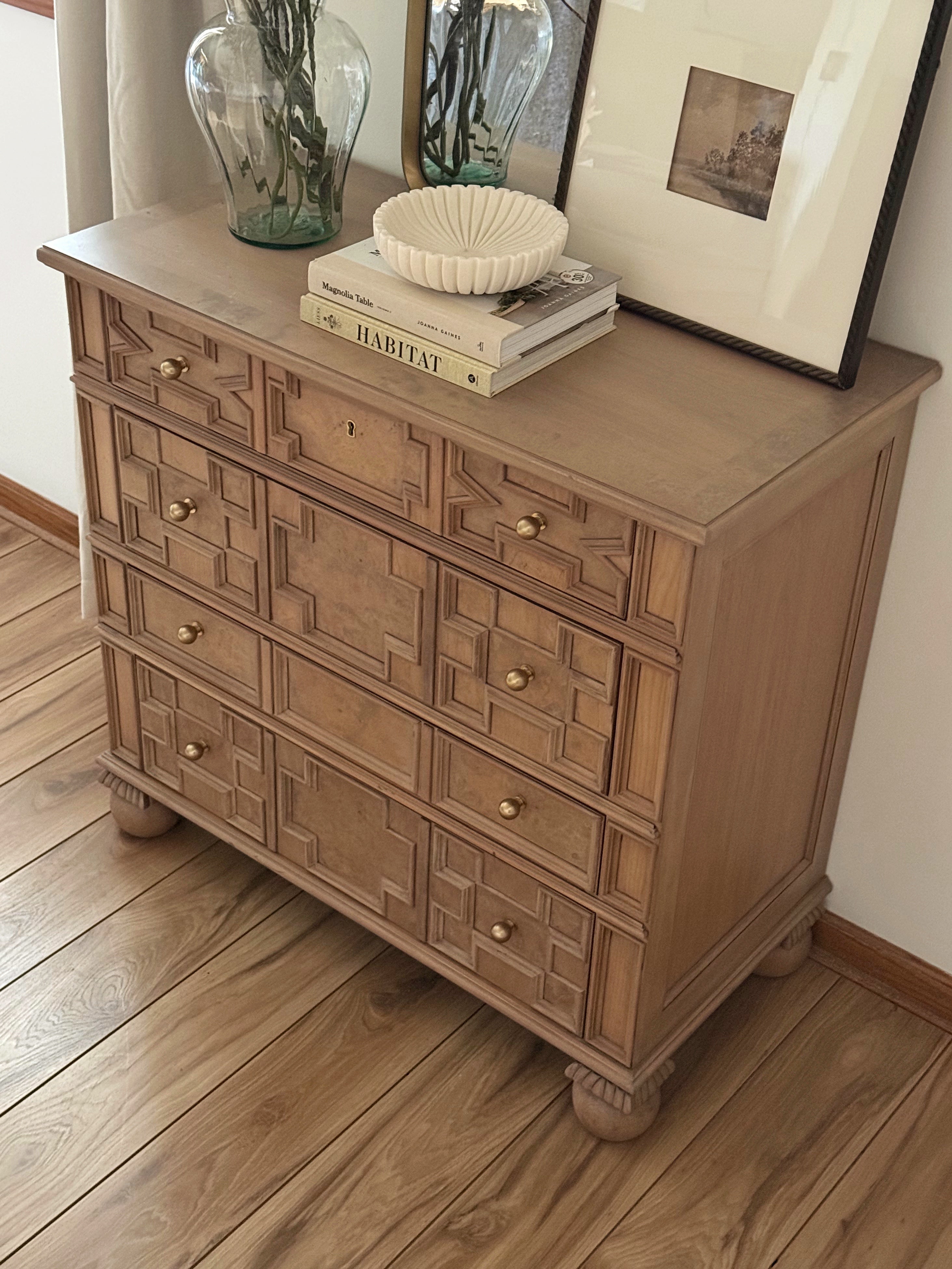 Vintage wood dresser with geometric carved drawer fronts and brass knobs, styled with neutral decor, books, and glass vases in a modern organic interior.